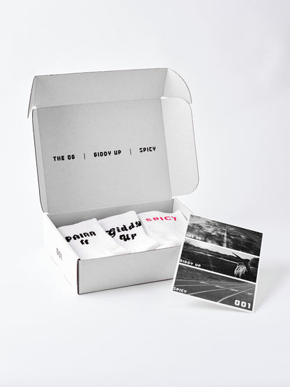 An open white cardboard box containing three white socks with black and pink text, alongside a black-and-white printed card, photographed on a clean white background.