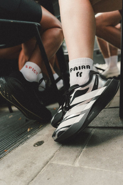 A close-up lifestyle shot of a person's legs and feet, showcasing OG PAIRR white crew socks with black and white striped running shoes. The setting is outdoors on a gray concrete pavement next to black metal chairs. SPICY white crew sock in on another person at the table in black shoes.