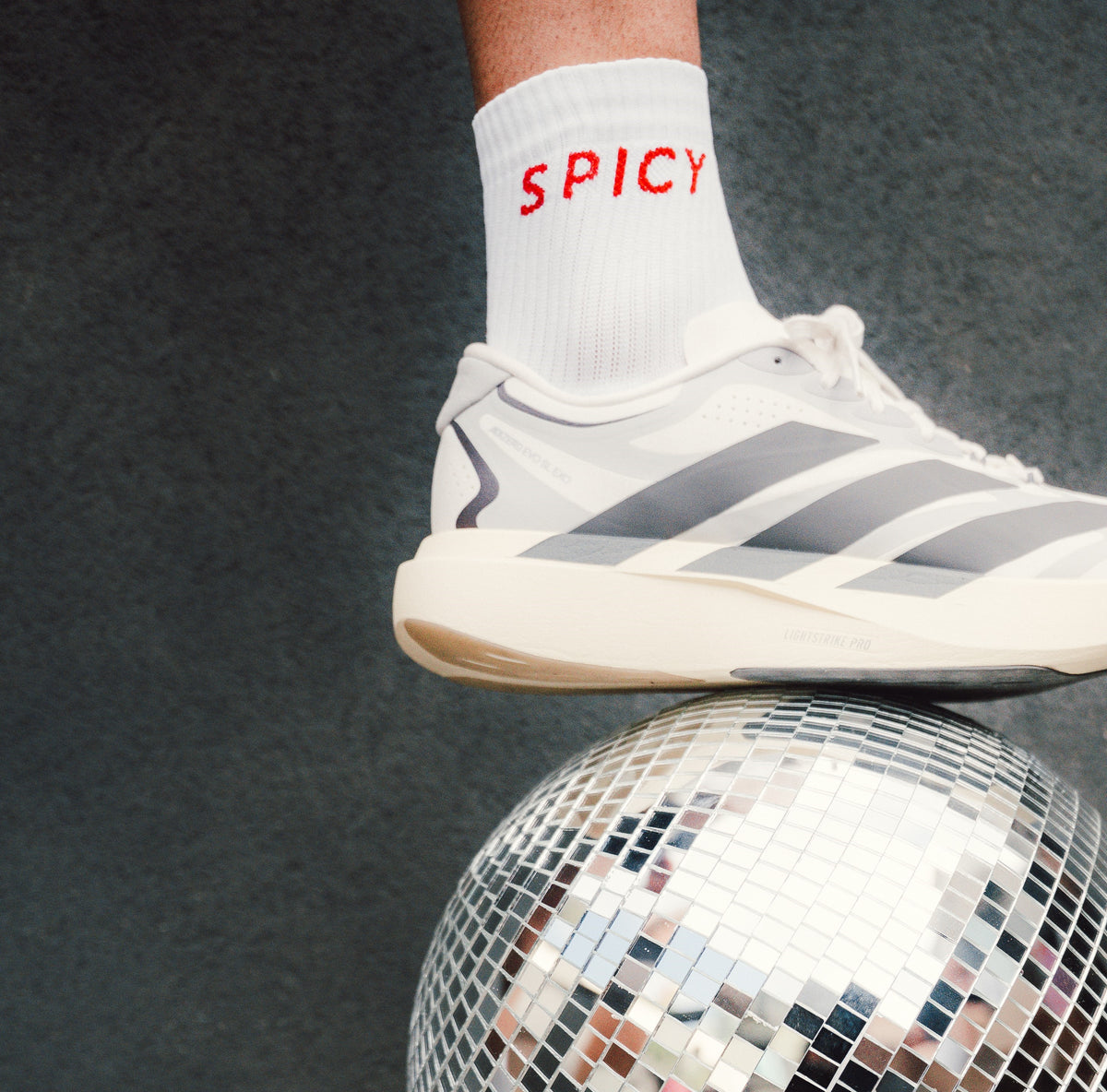 A close-up of a white Adidas sneaker with gray stripes, worn on a foot with a white sock printed with the word 'SPICY' in red, balancing on top of a mirrored disco ball against a dark gray background.