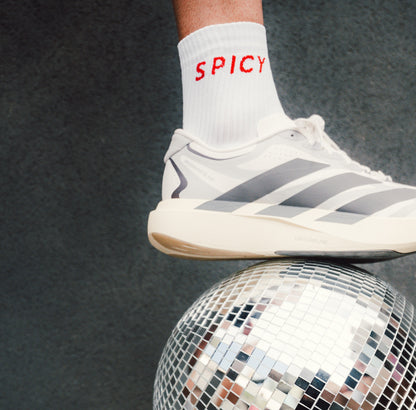 A close-up of a white Adidas sneaker with gray stripes, worn on a foot with a white sock printed with the word 'SPICY' in red, balancing on top of a mirrored disco ball against a dark gray background.